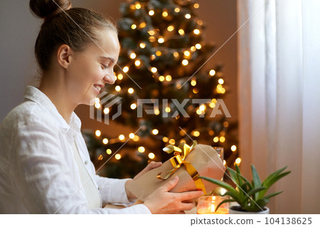 Indoor shot of smiling woman wearing white shirt sitting at table and holding christmas gift with gold bow on background of christmas tree with lights, expressing happiness and positive emotions. Indoor shot of smiling woman wearing white shirt sitting at table and holding christmas gift with gold bow on background of christmas tree with lights, expressing happiness and positive emotions. 104138625