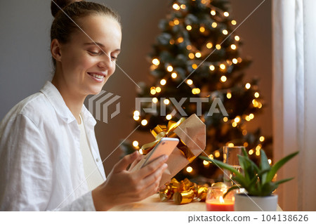 Side view portrait of young adult beautiful girl packing christmas gifts in decorated home interior near Christmas tree with bright lights, smiling happily, waiting for New Year holidays. Side view portrait of young adult beautiful girl packing christmas gifts in decorated home interior near Christmas tree with bright lights, smiling happily, waiting for New Year holidays. 104138626