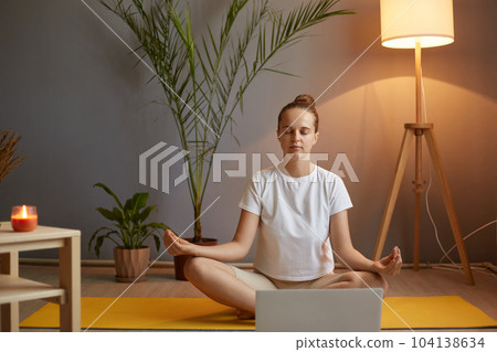 Indoor shot of relaxed calm sporty woman with hair bun wearing white t shirt practicing yoga at home in cozy room, sitting on mat in lotus pose with mudra gesture and closed eyes. Indoor shot of relaxed calm sporty woman with hair bun wearing white t shirt practicing yoga at home in cozy room, sitting on mat in lotus pose with mudra gesture and closed eyes. 104138634