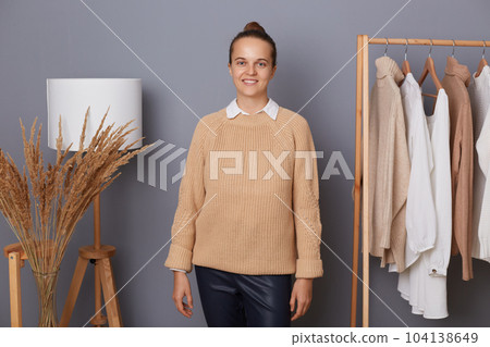 Portrait of attractive cheerful woman with bun hairstyle wearing beige sweater standing in fashion sore near clothing on hangers, looking at camera, presenting her online showroom. 104138649