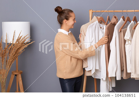 Young adult happy woman with bun hairstyle professional expert fashion stylist standing near clothes hang on shelf in designer store, selling trendy outfit in shop boutique. Young adult happy woman with bun hairstyle professional expert fashion stylist standing near clothes hang on shelf in designer store, selling trendy outfit in shop boutique. 104138651