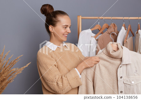 Attractive smiling woman with dark hair and bun hairstyle holding clothes on racks in modern boutique, choosing attire, trying to make stylish trendy look, expressing happiness. Attractive smiling woman with dark hair and bun hairstyle holding clothes on racks in modern boutique, choosing attire, trying to make stylish trendy look, expressing happiness. 104138656