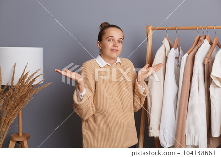 Uncertain confused woman wearing beige knitted sweater standing near shelves with clothes, looking with doubts, thinking and choosing trendy outfit, shopping in modern boutique, can`t make decision. 104138661
