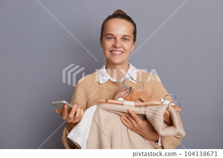 Attractive happy smiling positive woman shopper standing with clothing in hands and holding phone isolated over gray background, looking at camera with toothy smile, enjoying shopping. 104138671