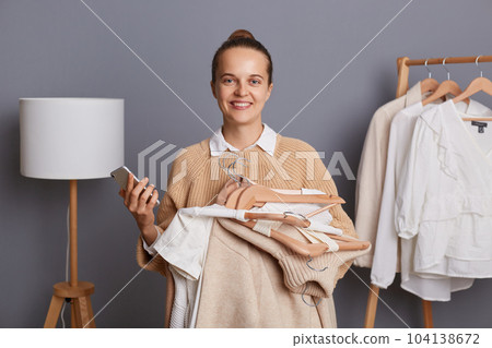Portrait of winsome pretty woman stylist holding clothes on racks in fashion store, holding mobile phone, using internet for searching new looks, looking at camera with positive expression. 104138672
