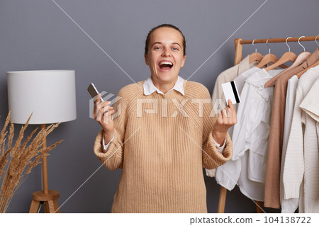 Indoor shot of excited amazed Caucasian woman holding credit card and smart phone in hands, paying online for her shopping, screaming happily, pays for new attire. Indoor shot of excited amazed Caucasian woman holding credit card and smart phone in hands, paying online for her shopping, screaming happily, pays for new attire. 104138722
