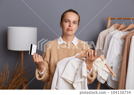 Indoor shot of sad upset woman holding clothing on hangers, cell phone and credit card, expressing sorrow and sadness, female shopper can't pay for her outfits. Indoor shot of sad upset woman holding clothing on hangers, cell phone and credit card, expressing sorrow and sadness, female shopper can't pay for her outfits. 104138734