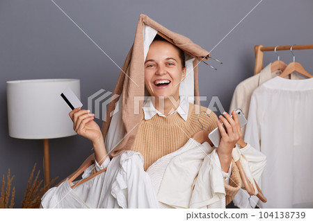 Image of smiling positive optimistic woman with clothes on her head holding clothing on hangers, doing shopping in mall, standing with credit card and smart phone. Image of smiling positive optimistic woman with clothes on her head holding clothing on hangers, doing shopping in mall, standing with credit card and smart phone. 104138739