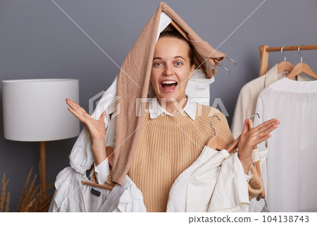 Photo of excited amazed extremely happy woman with clothes on her head holding clothing on hangers, standing with funny look, screaming happily, rejoicing to buy new attires. 104138743