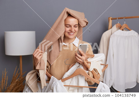 Portrait of delighted satisfied woman shopper with clothes on her head holding clothing on hangers, standing with smartphone in hands, looking at display with toothy smile. Portrait of delighted satisfied woman shopper with clothes on her head holding clothing on hangers, standing with smartphone in hands, looking at display with toothy smile. 104138749