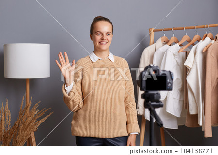 Horizontal shot of smiling beautiful Caucasian wearing beige jumper recording video in her showroon for her blog about fashion, waving hand, saying hello to followers. 104138771
