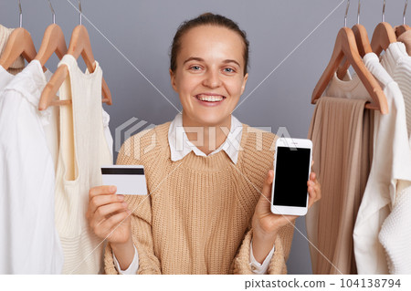 Indoor shot of smiling cheerful woman standing near clothes hang on shelf, showing mobile phone with empty display and credit card, presenting copy space for advertisement area. Indoor shot of smiling cheerful woman standing near clothes hang on shelf, showing mobile phone with empty display and credit card, presenting copy space for advertisement area. 104138794