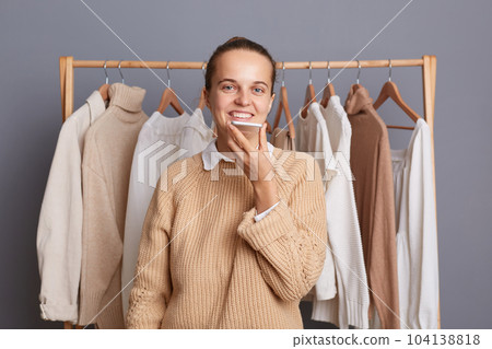 Indoor shot of attractive beautiful woman standing near hanger rack with attires in fashion store, holding mobile phone and recording voice message, looking at camera, posing in showroom. Indoor shot of attractive beautiful woman standing near hanger rack with attires in fashion store, holding mobile phone and recording voice message, looking at camera, posing in showroom. 104138818