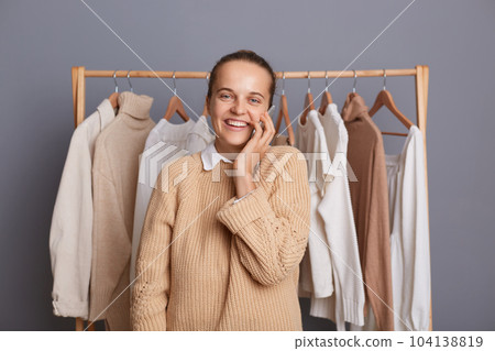 Indoor shot of attractive joyful happy Caucasian woman standing near clothes hang on shelf and talking on smart phone with her new client or buyer, looking at camera. Indoor shot of attractive joyful happy Caucasian woman standing near clothes hang on shelf and talking on smart phone with her new client or buyer, looking at camera. 104138819