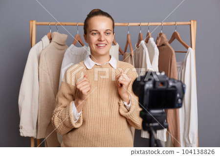 Portrait of woman blogger or designer with bun hairstyle wearing beige jumper standing against gray wall with clothes on hangers on shelf, recording video, telling about fashion trends. 104138826