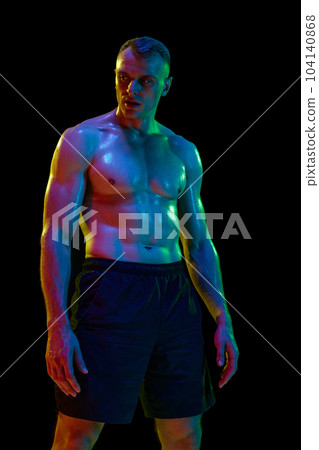 Hard training. Handsome man with muscular, relief, fit, shirtless body posing against black studio background in neon light 104140868