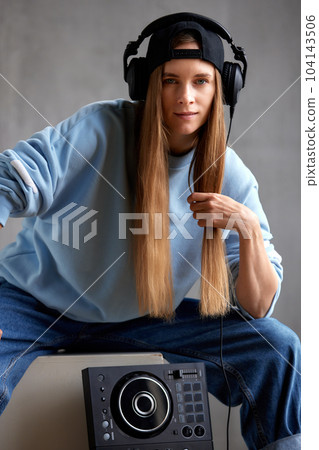 A young pretty long-haired DJ girl in a blue sweater, jeans, a black baseball cap and black headphones sits with a DJ mixing console. Studio shot, gray background. A young pretty long-haired DJ girl in a blue sweater, jeans, a black baseball cap and black headphones sits with a DJ mixing console. Studio shot, gray background. 104143506