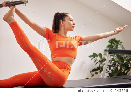 A young girl is doing Pilates on a reformer bed in a bright studio. A slender brunette in an orange bodysuit is doing exercises for flexibility of the body. The concept of a healthy lifestyle. 104143516