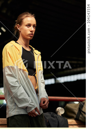 Young fitness girl getting ready for a workout at the stadium. Attractive slender blonde in a black top and a yellow-gray sports jacket is focused on training. Active lifestyle. Half-length shot. 104143554