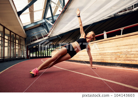 A young athlete girl is warming up before training at the stadium. A slender blonde woman in a black tracksuit does a plank exercise to strengthen her muscles. Sports and recreation. A young athlete girl is warming up before training at the stadium. A slender blonde woman in a black tracksuit does a plank exercise to strengthen her muscles. Sports and recreation. 104143603