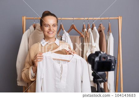 Indoor shot of pretty pleased positive woman with bun hairstyle recording video on camera, holding white shirt on hanger, showing fashionable look for her female followers. 104143683