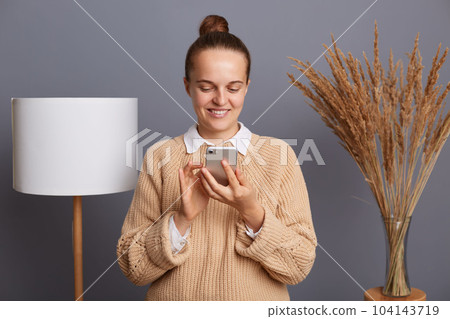 Image of joyful woman wearing warm sweater using cell phone for checking cocial networks, expressing positive emotions, standing against gray wall with lamp and dried flower on background. 104143719