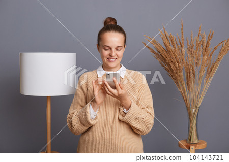 Horizontal shot of smiling joyful woman wearing beige warm sweater standing indoor near lamp in living room, holding mobile phone, checking social networks, reading messages, typing. Horizontal shot of smiling joyful woman wearing beige warm sweater standing indoor near lamp in living room, holding mobile phone, checking social networks, reading messages, typing. 104143721