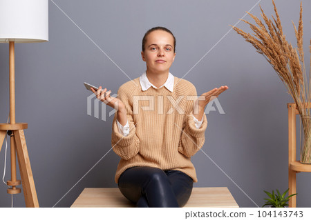 I don't know. Attractive confused woman wearing beige sweater sitting on chair with raised arms and looking at camera, holding mobile in hands, doesn't know answer. I don't know. Attractive confused woman wearing beige sweater sitting on chair with raised arms and looking at camera, holding mobile in hands, doesn't know answer. 104143743