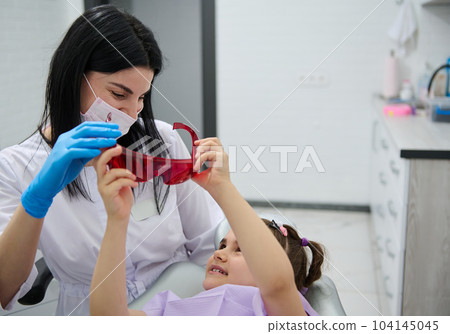 Pediatric dentistry. Dental practice. Pleasant female dentist doctor talking to a little patient before dental check up. Oral health and hygiene. The concept of baby teeth treatment. Caries prevention 104145045