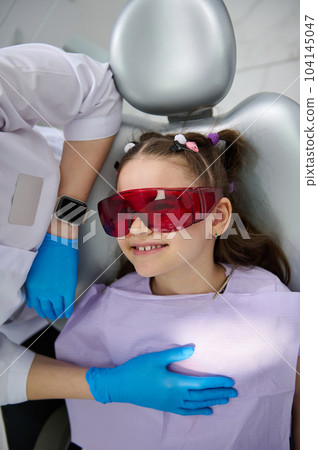 Close-up top view of a smiling happy little child girl, patient in UV goggles, visiting dentist in pediatric dentistry clinic. Dental health and oral hygiene. The concept of baby teeth treatment 104145047