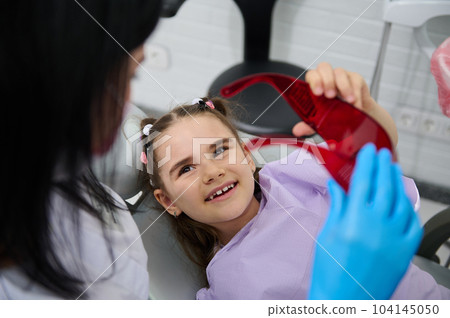 Smiling kid girl visiting dentist, curing tooth, putting braces in modern pediatric dentistry clinic. Female dentist, dental hygienist treating caries to little child patient. Oral health and hygiene 104145050