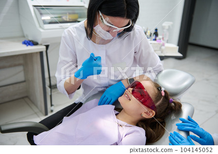 Portrait of beautiful child 6 years old, adorable little girl with open mouth, being examined by a dental hygienist, orthodontist while sitting in dentist chair in pediatric dentistry clinic. Close-up 104145052