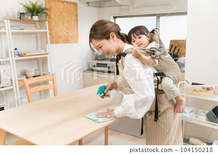 A young woman wiping the table while carrying a child on her back with a shoulder strap in the dining room A young woman wiping the table while carrying a child on her back with a shoulder strap in the dining room 104150218