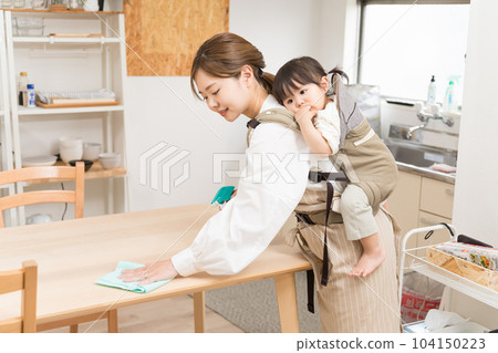 A young woman wiping the table while carrying a child on her back with a shoulder strap in the dining room 104150223