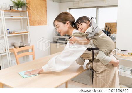 A young woman wiping the table while carrying a child on her back with a shoulder strap in the dining room 104150225
