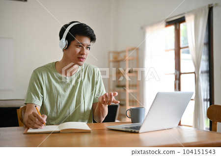 A smart Asian male college student wears headphones and focuses on studying an online 104151543