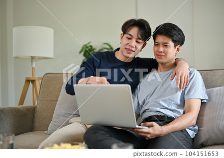 A happy Asian gay man enjoys watching a movie through a laptop with his boyfriend on a couch A happy Asian gay man enjoys watching a movie through a laptop with his boyfriend on a couch 104151653