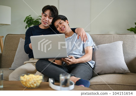 A happy Asian gay man is hugging his boyfriend while watching a movie on a couch together. A happy Asian gay man is hugging his boyfriend while watching a movie on a couch together. 104151654