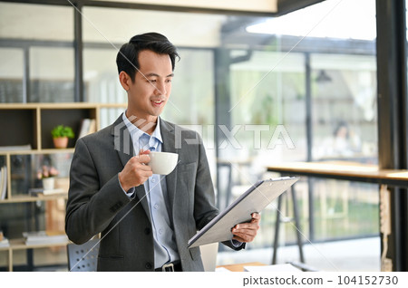 A handsome Asian businessman reading business report and sipping coffee in the office. A handsome Asian businessman reading business report and sipping coffee in the office. 104152730