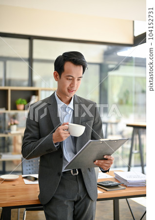 A smart Asian businessman reading document and sipping coffee while standing in the office. A smart Asian businessman reading document and sipping coffee while standing in the office. 104152731