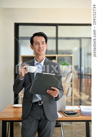 A successful businessman stands in his office with a cup of coffee and a document folder in his hand A successful businessman stands in his office with a cup of coffee and a document folder in his hand 104152734