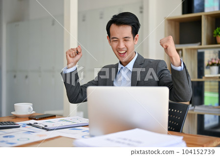 A cheerful Asian businessman looking at his laptop screen, and celebrating his success in the office. 104152739