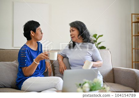Two mature middle-aged Asian women are enjoying their morning coffee and talking on a sofa Two mature middle-aged Asian women are enjoying their morning coffee and talking on a sofa 104154789