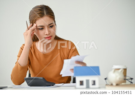 A stressed Asian woman sits at a table, checking her bills, calculating monthly expenses 104155451
