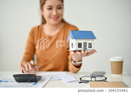A female real estate agent holding and looking at a house model at her desk. A female real estate agent holding and looking at a house model at her desk. 104155461