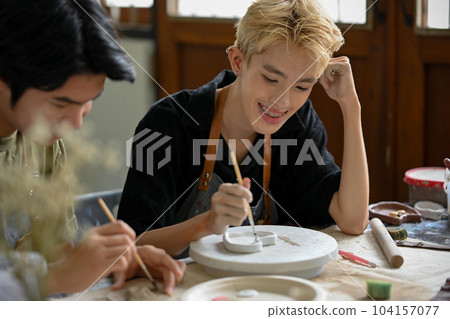 A happy Asian gay man enjoys painting a ceramic plate in a workshop with his boyfriend. 104157077