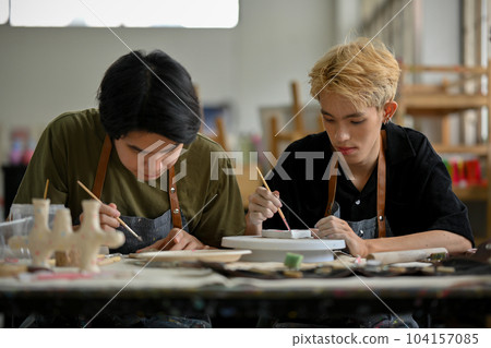A young Asian gay man focuses painting a ceramic plate in a creative handicraft workshop 104157085