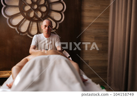 Portrait of focused professional male masseur using massage oil for deep muscle relaxation. Unrecognizable man client enjoying being gently massaged lying on table in luxury spa salon. Portrait of focused professional male masseur using massage oil for deep muscle relaxation. Unrecognizable man client enjoying being gently massaged lying on table in luxury spa salon. 104158749