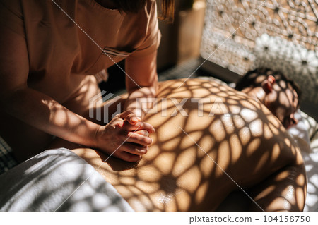 Close-up cropped shot of unrecognizable female masseuse giving massage to unrecognizable man in luxury spa salon lying on massage table in sunlight and beautiful shadows. Concept of healthy lifestyle. 104158750
