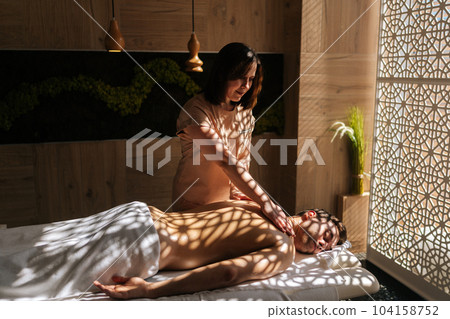 Side view of professional female masseuse massaging shoulder blades of muscular sportsman lying on massage table in luxury wellness center. Male client came to massage therapy due to body aches. 104158752
